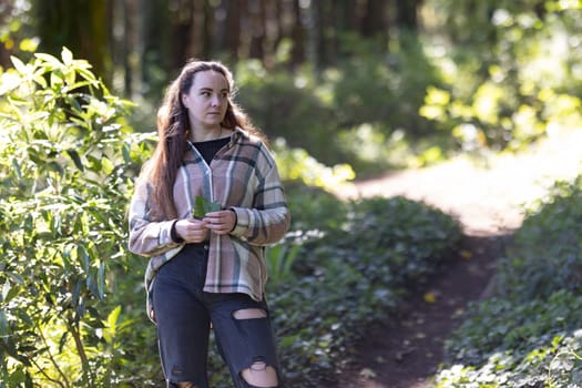 A woman is walking along a trail in a wooded area. Surrounded by tall trees and greenery, she walks with purpose carrying a backpack.