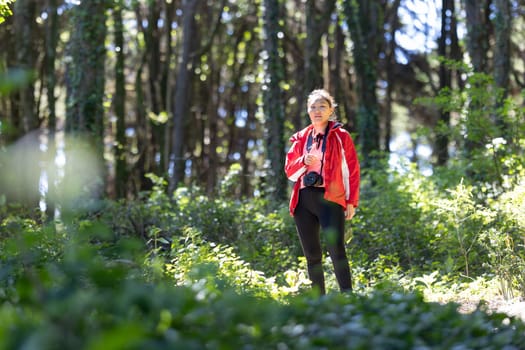 A woman wearing a red jacket walks through a dense forest, surrounded by tall trees and fallen leaves. The forest path is visible ahead, with sunlight filtering through the branches.