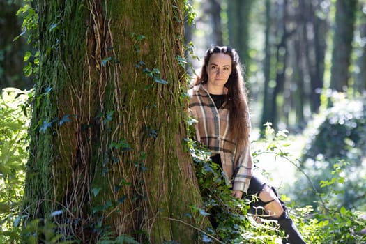 A woman is seated next to a tree in a dense forest, surrounded by lush greenery. She appears relaxed, enjoying the peaceful atmosphere while spending time outdoors with friends.