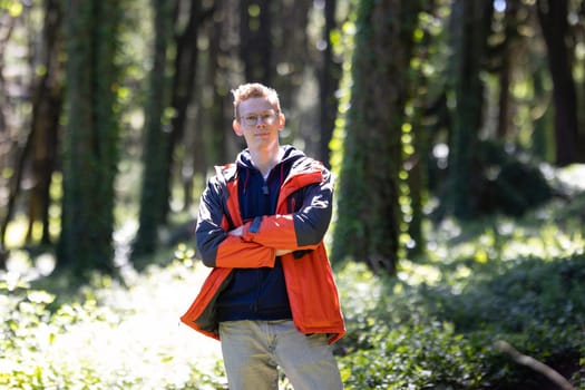 A man standing with his arms crossed in the midst of a dense forest, surrounded by tall trees and underbrush. He appears contemplative and deep in thought as he gazes into the distance.