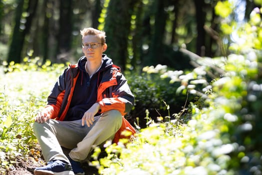 A young boy is seated on a large rock amongst the trees in a wooded area. He appears relaxed and contemplative, taking in his surroundings.