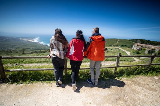 Three friends stand together on a hill, gazing out at the vast expanse of the ocean spread out before them. They appear relaxed, taking in the view and enjoying each others company on a clear day.