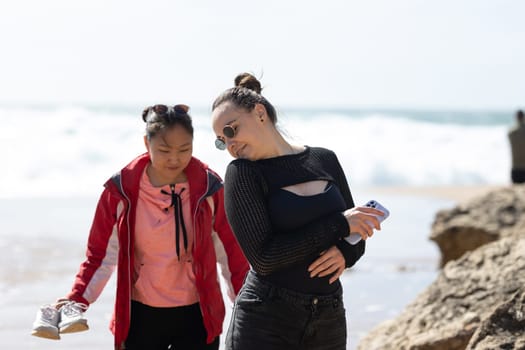 A woman and a young girl are walking together on the sandy beach, enjoying the fresh air and the sound of the crashing waves. The ocean stretches endlessly in the background.
