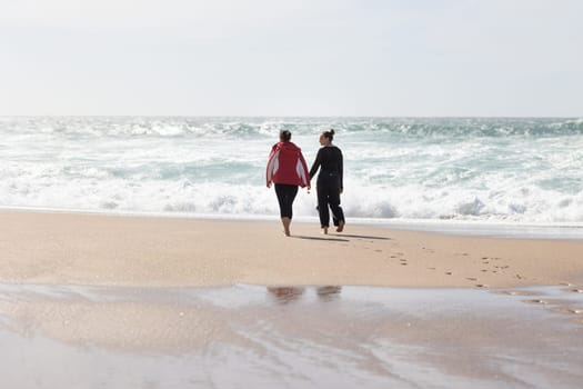 A pair of individuals, likely friends, strolling along the sandy beach while interlocking hands. The two figures are engaged in a shared activity, enjoying the seaside atmosphere.