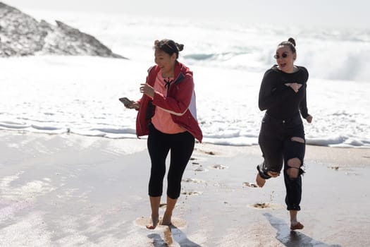 Two women, friends, are seen energetically running along a sandy beach with crashing waves from the ocean in the background. The women appear determined and focused as they enjoy their outdoor exercise session.