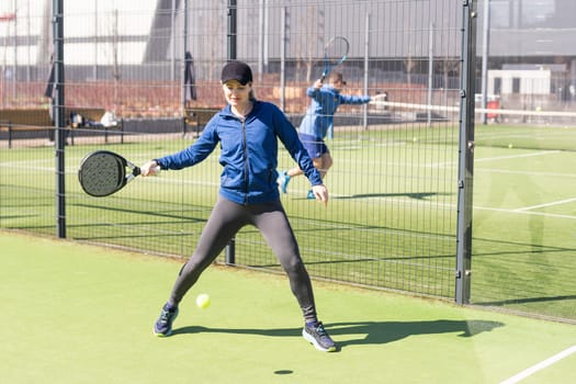 A girl in sportswear is training on a paddle tennis court. The girl is hitting the ball against the glass to make a rebound. Concept of women playing paddle. High quality photo