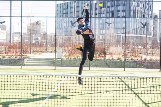 A padel player jump to the ball, good looking for posts and poster. Man with black racket playing a match in the open behind the net court outdoors. Professional sport concept with space for text. High quality photo
