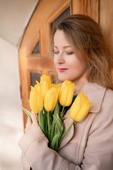 A woman is holding a bouquet of yellow tulips. She is wearing a tan coat and a green shirt