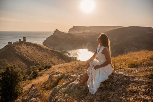 A woman in a white dress sits on a rock overlooking a body of water. The scene is serene and peaceful, with the woman enjoying the view and the calmness of the surroundings