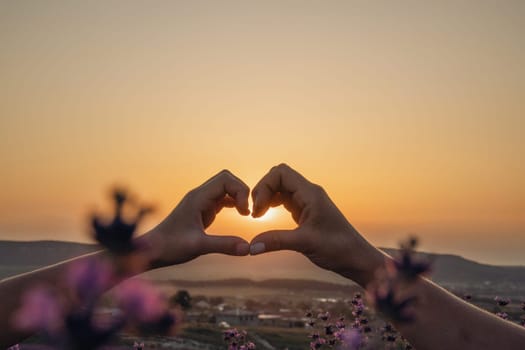 Two hands forming a heart shape in front of a purple field. The sun is setting in the background, creating a warm and romantic atmosphere