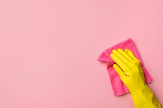 Employee hand in yellow rubber protective glove hold red or pink rag wiping pink wall background. House cleaning service, general or regular cleanup concept. Empty place for text or design