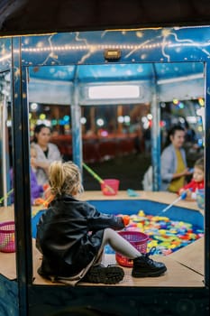 Little girl catches fish with a toy fishing rod while sitting by a toy pool at a fair with children. Back view. High quality photo