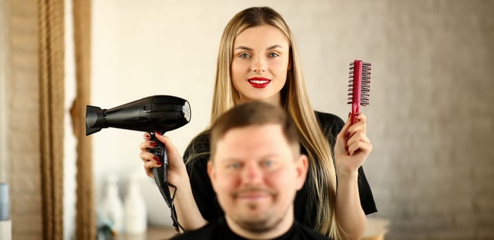 Blonde Hairstylist Using Dryer and Comb for Man.Young Hairdresser Styling Male Haircut with Hairdryer and Hairbrush. Woman Stylist Holding Tool for Hairstyling. Beautician and Client Looking at Camera