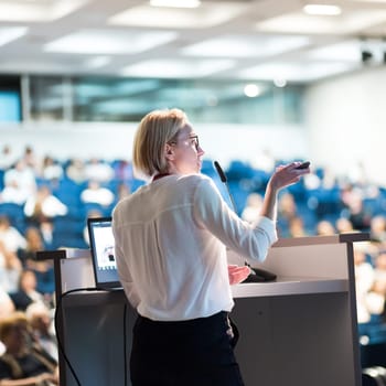 Female speaker giving a talk on corporate business conference. Unrecognizable people in audience at conference hall. Business and Entrepreneurship event