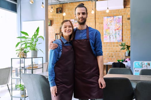 Small business team portrait of hugging of successful colleagues partners young man woman in aprons posing looking at camera at workplace in restaurant coffee shop cafeteria. Partnership teamwork work
