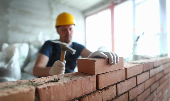 Portrait of skilled man constructing big concrete wall and using special hammer to properly fulfill task to gently lay bricks on unfinished structure. Building concept