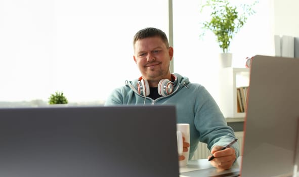 Handsome smiling male student using online education service and drink tea or coffee. Young man looking in laptop display watching training course and listening it with headphones.