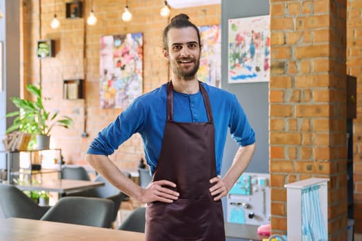 Confident successful young man service worker owner in apron, handsome male looking at camera in restaurant cafeteria coffee pastry shop interior. Small business staff occupation entrepreneur work
