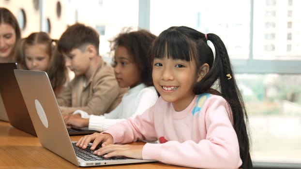 Asian girl working on laptop and looking at camera. Diverse children coding or programing engineering code or prompt by using computer in STEM technology class while teacher explain system. Erudition.