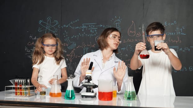 Caucasian boy mixing chemical liquid while teacher giving advice. Professional instructor wearing lab suit looking for diverse student at table with beaker filled with colored solution. Erudition.