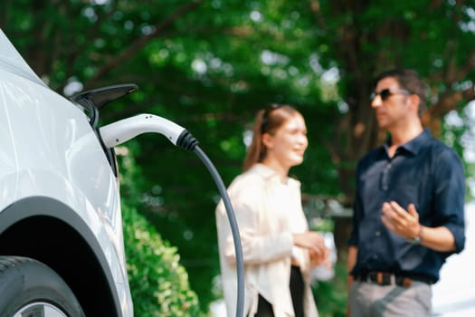 Young couple recharge electric car battery from charging station in green city park in springtime. Rechargeable EV car for sustainable environmental friendly urban travel lifestyle. Panorama Expedient