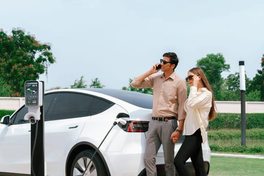 Young couple recharge her EV electric vehicle at green city park parking lot while talking on phone. Sustainable urban lifestyle for environment friendly EV car with battery charging station.Expedient