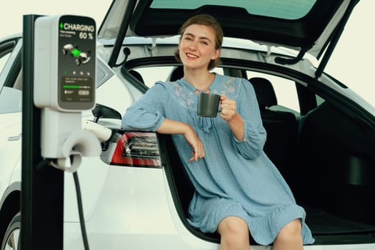 Holiday road trip vacation with young woman sitting in the car trunk with coffee while recharging electric vehicle with alternative energy. Environmental friendly travel wit EV car. Perpetual