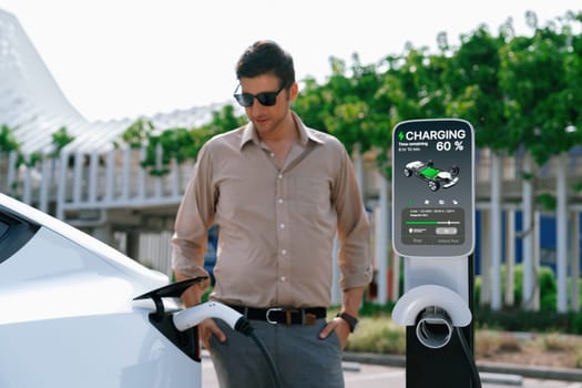 Young man put EV charger to recharge electric car's battery from charging station in city commercial parking lot. Rechargeable EV car for sustainable environmental friendly urban travel. Expedient