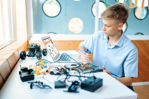 Blonde hair schoolboy in blue shirt watching code on laptop and use screwdriver fix motherboard in STEM class. Around laptop put controller, electric wire, battery charger and laptop. Edification.