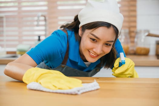 Professional Asian cleaner in yellow gloves wipes kitchen counter with liquid spray. Housekeeping service emphasizes home cleanliness and hygiene. Clean disinfect home care. maid household job.
