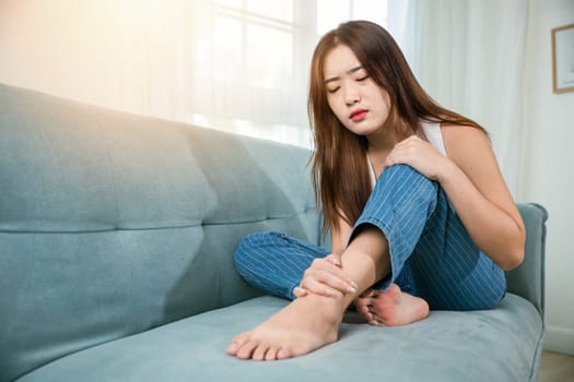 Asian young woman sitting on sofa holding her feet and stretch muscles have symptoms feeling pain, beautiful female problems with foot at home, painful ankle injury, Health care and medical concept