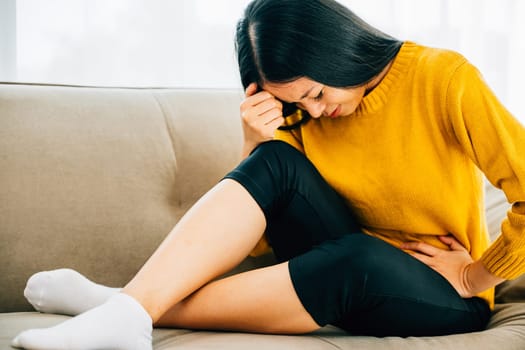 A woman of Asian descent suffers stomach ache on a living room couch at home. Depicting discomfort symptoms and the importance of medical care for stomachache. Health care abdomen bloating concept