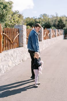 Little girl looks back while walking with her dad holding hands along the road along the fence. Back view. High quality photo