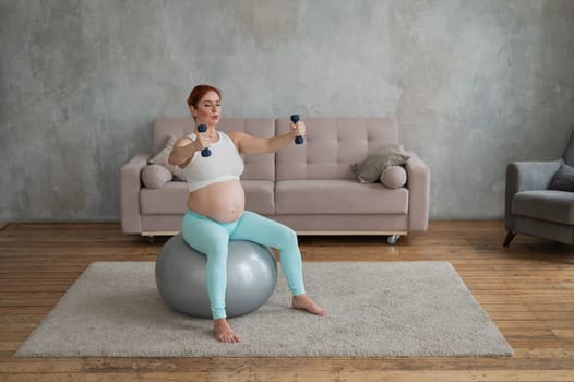 Pregnant woman doing exercises with dumbbells while sitting on a fitness ball at home