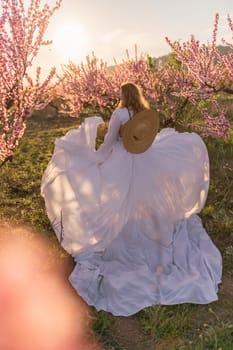 Woman blooming peach orchard. Against the backdrop of a picturesque peach orchard, a woman in a long white dress and hat enjoys a peaceful walk in the park, surrounded by the beauty of nature