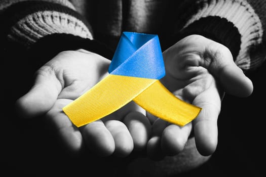 childs hand holds blue and yellow ukraine ribbon on dark background. black and white color. concept needs help and support, truth will win