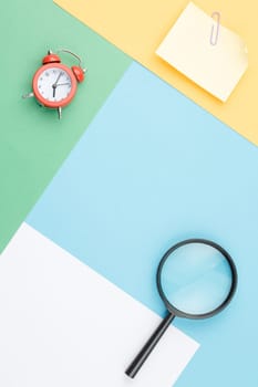 Colored paper background of geometric shapes with magnifying glass, alarm clock, note paper with paper clip and flower in the pot Office composition. Flat lay