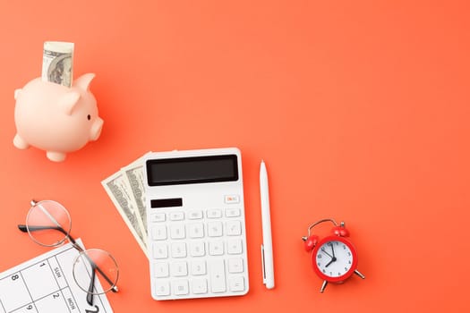 White calculator with pig piggy bank and dollars, pen, glasses, calendar sheet with dates alarm clock on orange isolated background flat lay top view.