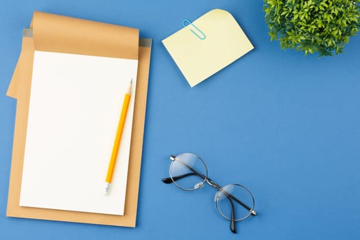 Open notebook with white blank sheet, glasses, pencil, note sheet with paper clip and houseplant with green leaves on blue isolated background. Top view.