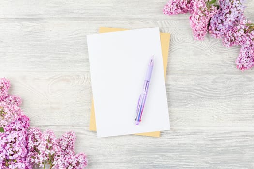 Craft sheets of paper with pen and lilac flowers on a white wooden background. Top view.