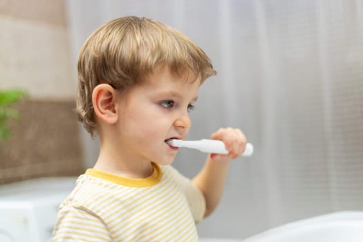 Boy with an electric toothbrush in the bathroom. Daily dental care concept. Side view portrait with copy space for health education and advertisements.