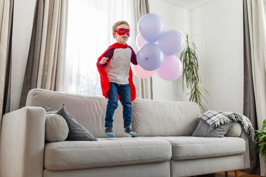 Child in superhero costume with balloons on couch. Indoor casual studio portrait with copy space. Imagination and play concept. Design for banner, poster, invitation card.