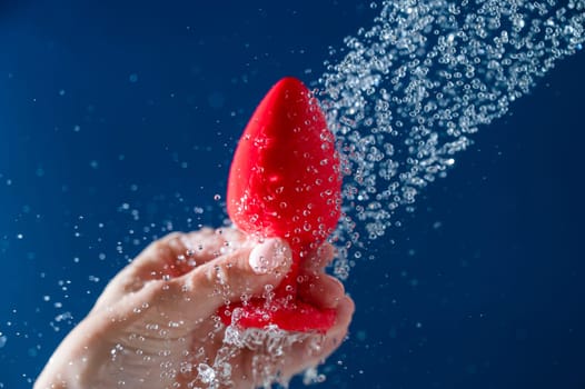 Woman washing red anal plug under shower on blue background