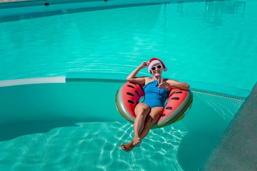 A happy woman in a blue bikini, a red and white Santa hat and sunglasses poses in the pool in an inflatable circle with a watermelon pattern, holding a glass of champagne in her hands. Christmas holidays concept