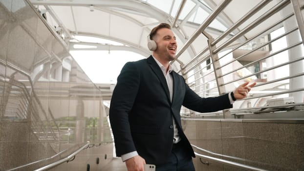 Business man dance to relaxing music from headphone at modern corridor. Cheerful project manager wear headset and moving gesture to celebrate successful song with lively mood in urban city. Urbane.