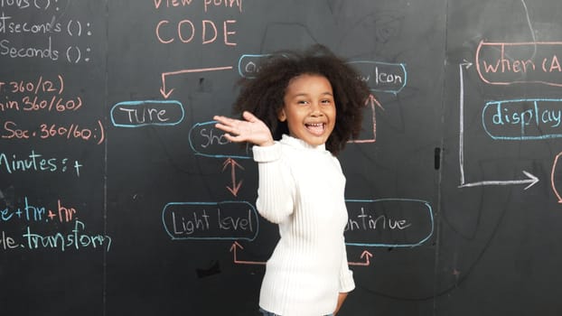 Happy african girl writing blackboard and turnaround to waving at camera while standing at board with engineering prompt or coding, programing system written in STEM technology classroom. Erudition.