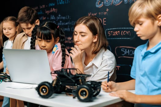 Skilled kids learning to program remote car. Schoolgirl in pink cloth use laptop for coding. Other child watch her code while teacher watch her. motherboard and electric wire also on table. Erudition.