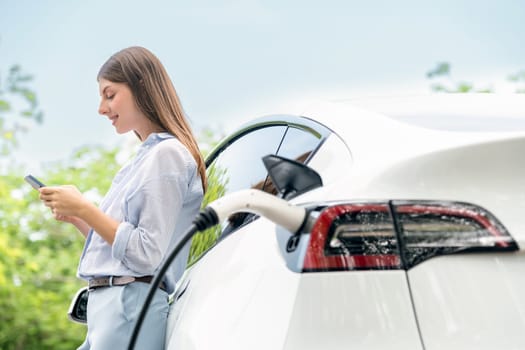 Young woman using smartphone online banking application to pay for electric car battery charging from EV charging station during vacation holiday road trip at national park or summer forest. Exalt