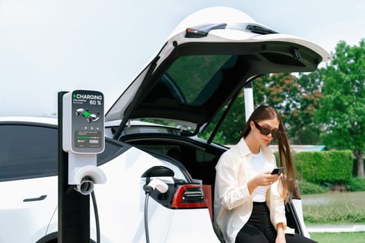 Young woman use smartphone to pay for electricity at public EV car charging station green city park. Modern environmental and sustainable urban lifestyle with EV vehicle. Expedient