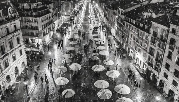 A black and white photo of a city street with many umbrellas and people. Scene is lively and bustling, with people walking around and enjoying the night. The umbrellas provide a sense of shelter